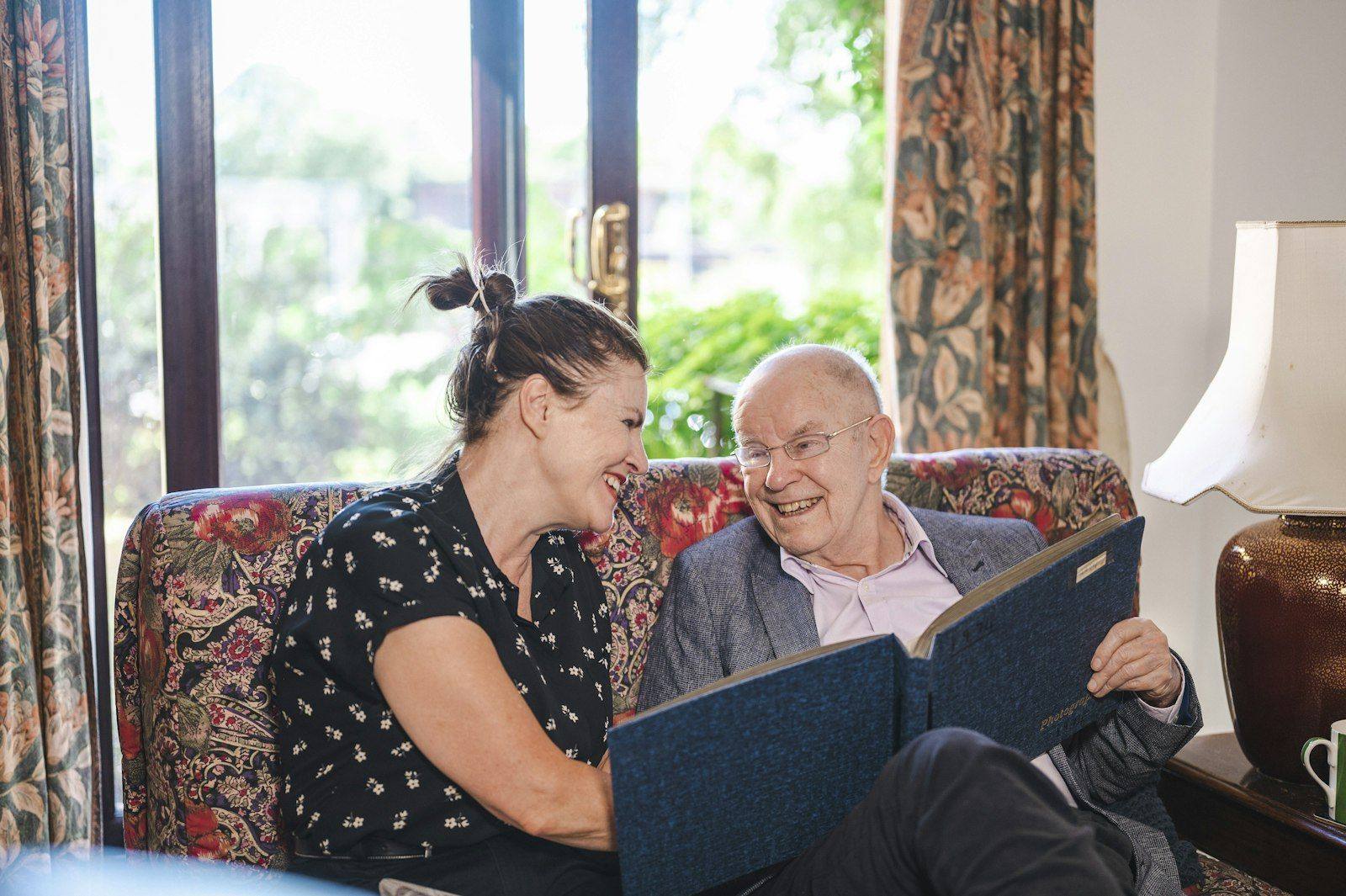 An older man and his carer smiling over a photo album together in his own front room.