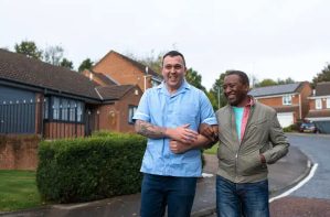 A carer and an older man walking arm in arm outdoors.